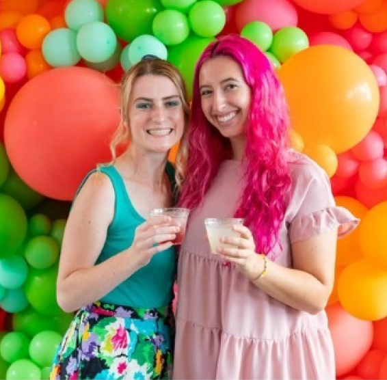 A smiling Rebecca Neckritz and Christina Mitchell pose closely to each other while holding colorful beverages.